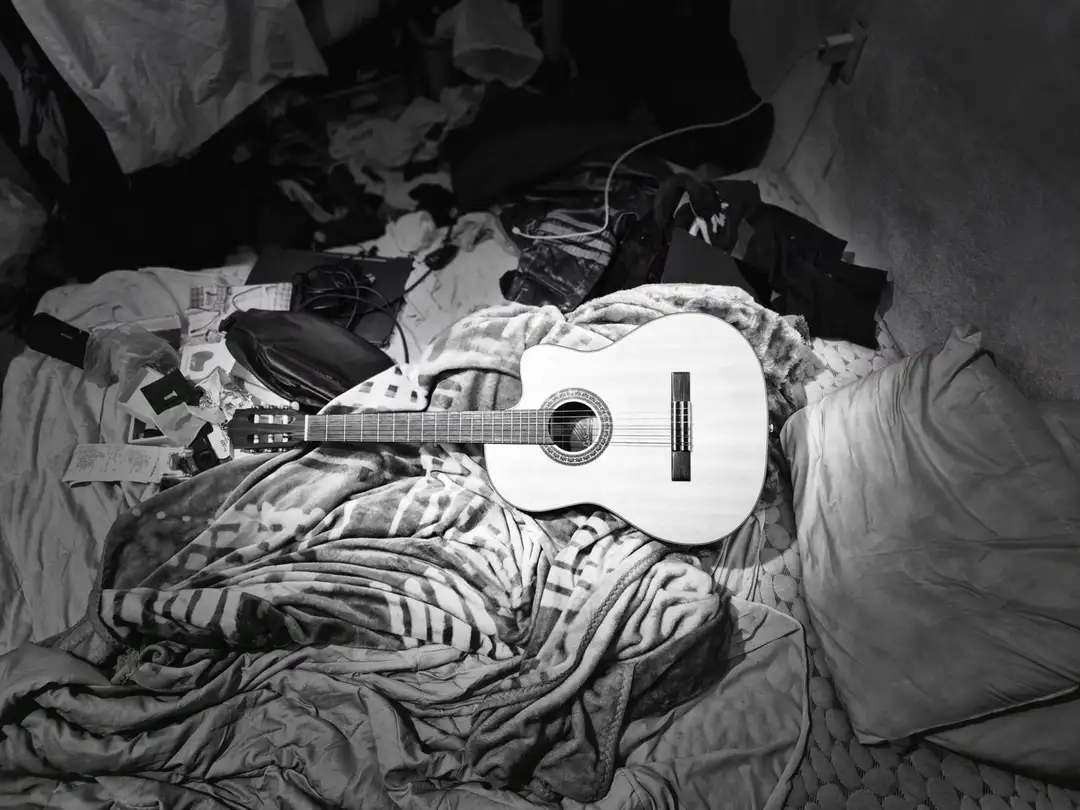 Guitarra clásica reposando sobre una cama desordenada en blanco y negro, símbolo de la honestidad y la intimidad del proceso musical.